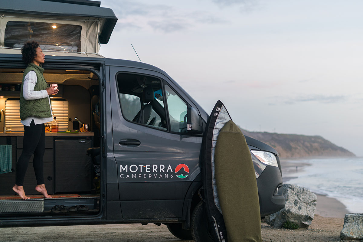 Woman Viewing Ocean and Drinking Coffee from Moterra California Campervan Rental with Surfboards